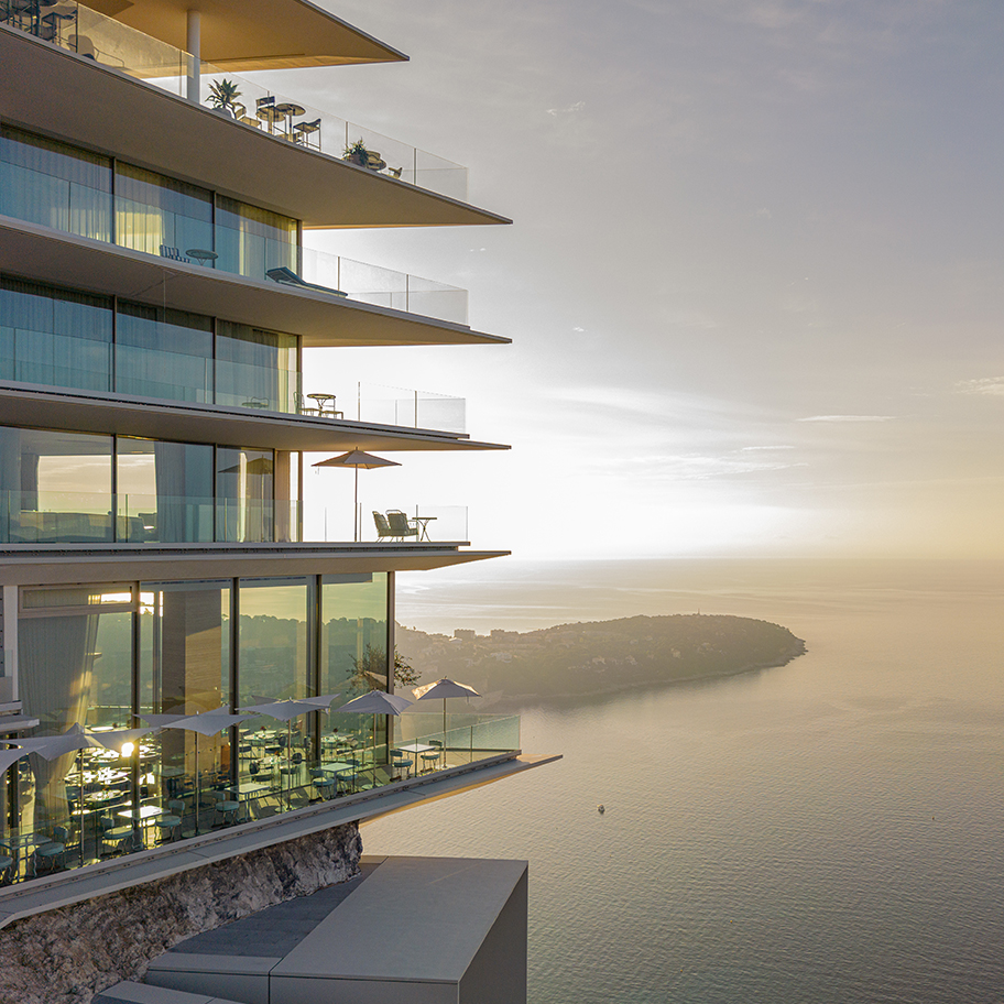 View of the side of The Maybourne Riviera hotel with Riviera Restaurant on the ground floor, rooms and suites above, and the Riviera landscape with the sea in the background.