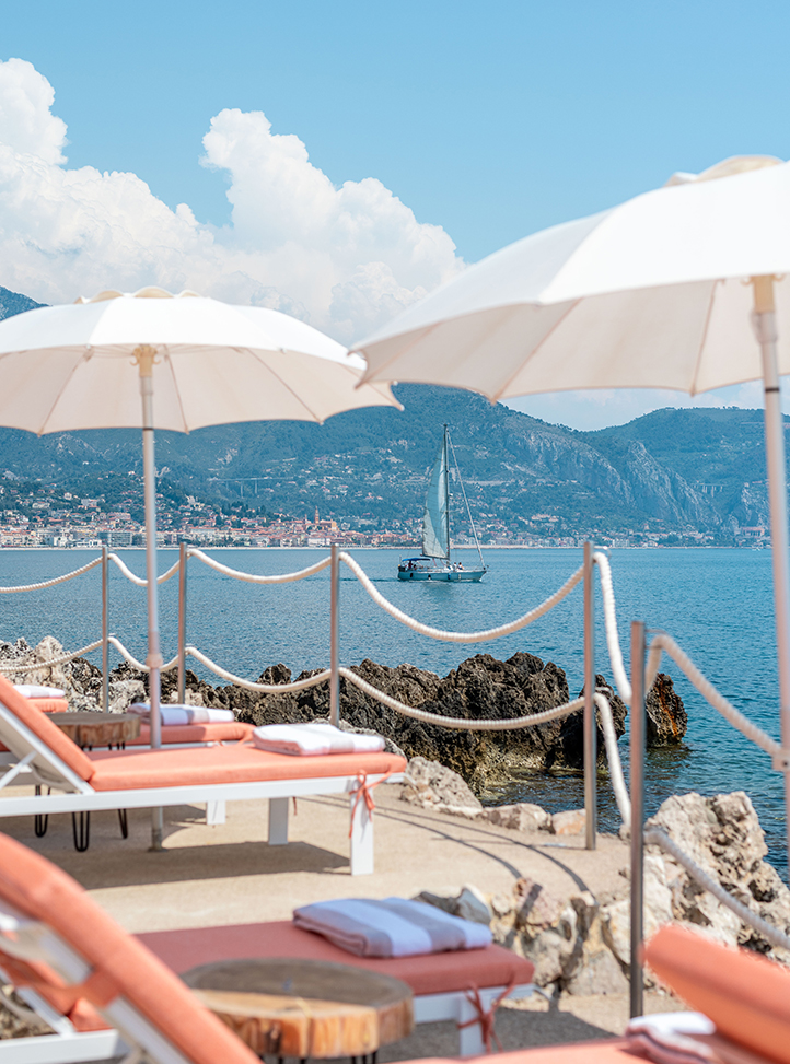 Les transats corail de Maybourne La Plage sont sous des parasols blanc; et donnent sur la mer avec un voilier et la côte en arrière-plan.