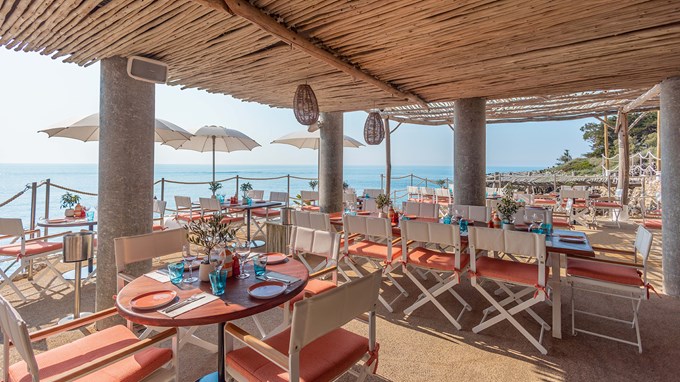 Maybourne La Plage dining area with round wooden tables, white and pink chairs and glassware on the table. There is a view on the sea in the background.
